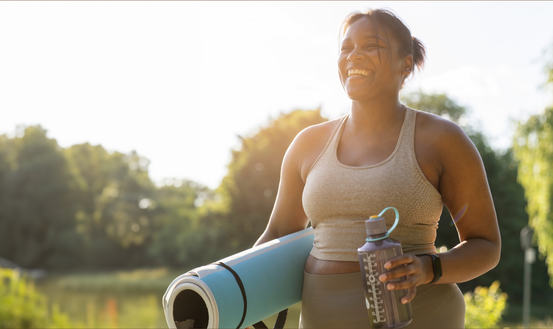 Woman smiling outdoors holding a yoga mat and water bottle